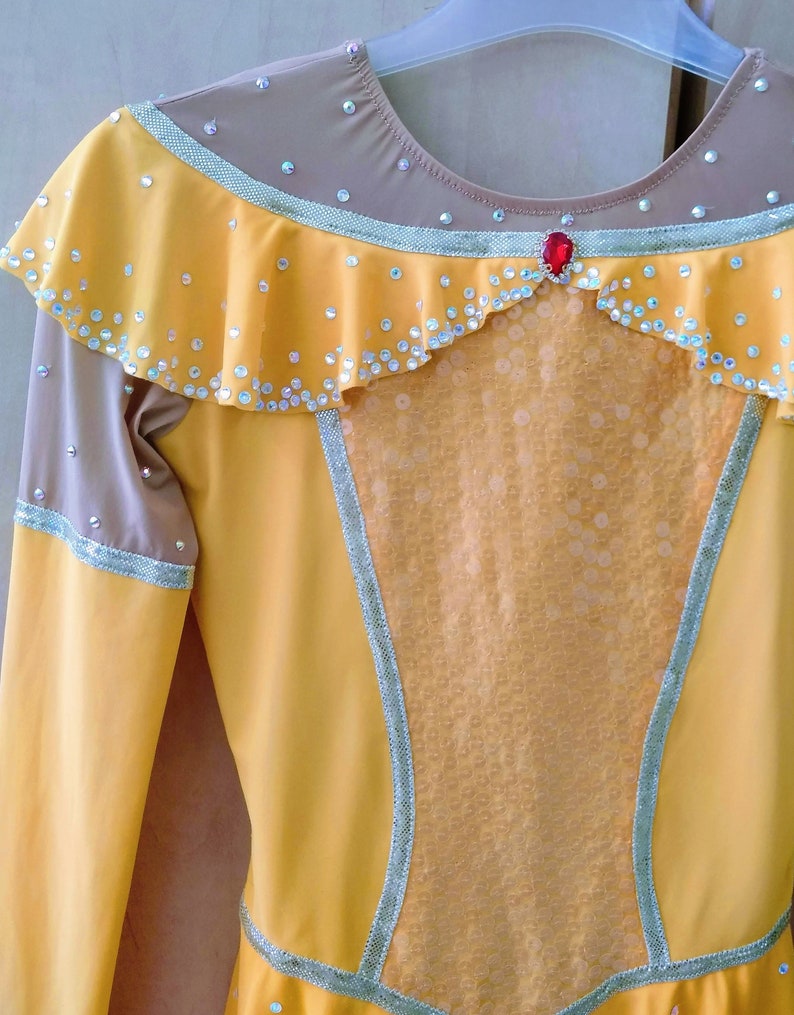 K&ouml;nnte beinhalten: Ein gelbes und braunes Eiskunstlaufkleid mit einem R&uuml;schenkragen, einem roten Strassstein und einem durchsichtigen Einsatz in der Mitte.