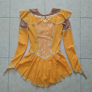 K&ouml;nnte beinhalten: Ein gelbes Eiskunstlaufkleid mit langen &Auml;rmeln und einem R&uuml;schenkragen. Das Kleid ist mit Pailletten und Strasssteinen verziert.