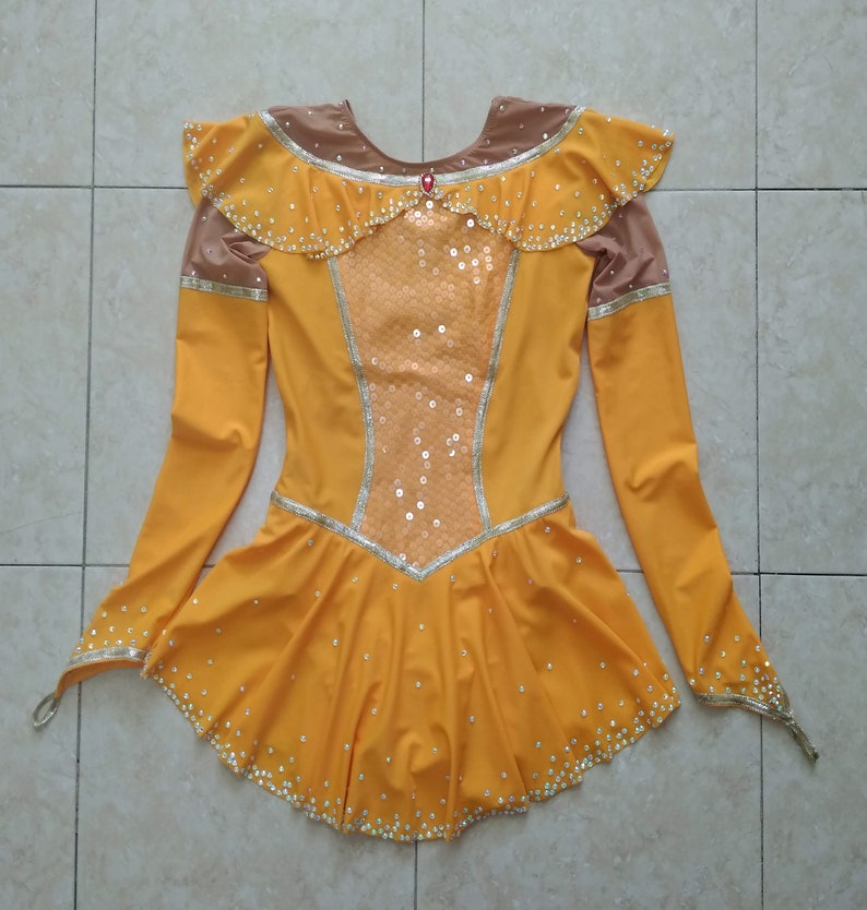 K&ouml;nnte beinhalten: Ein gelbes Eiskunstlaufkleid mit langen &Auml;rmeln und einem R&uuml;schenkragen. Das Kleid ist mit Pailletten und Strasssteinen verziert.