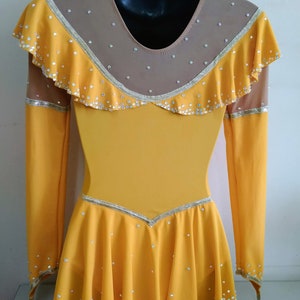K&ouml;nnte beinhalten: Ein gelbes Eiskunstlaufkleid mit langen &Auml;rmeln und einem R&uuml;schenkragen. Das Kleid ist mit Strasssteinen und einem goldenen Besatz verziert.