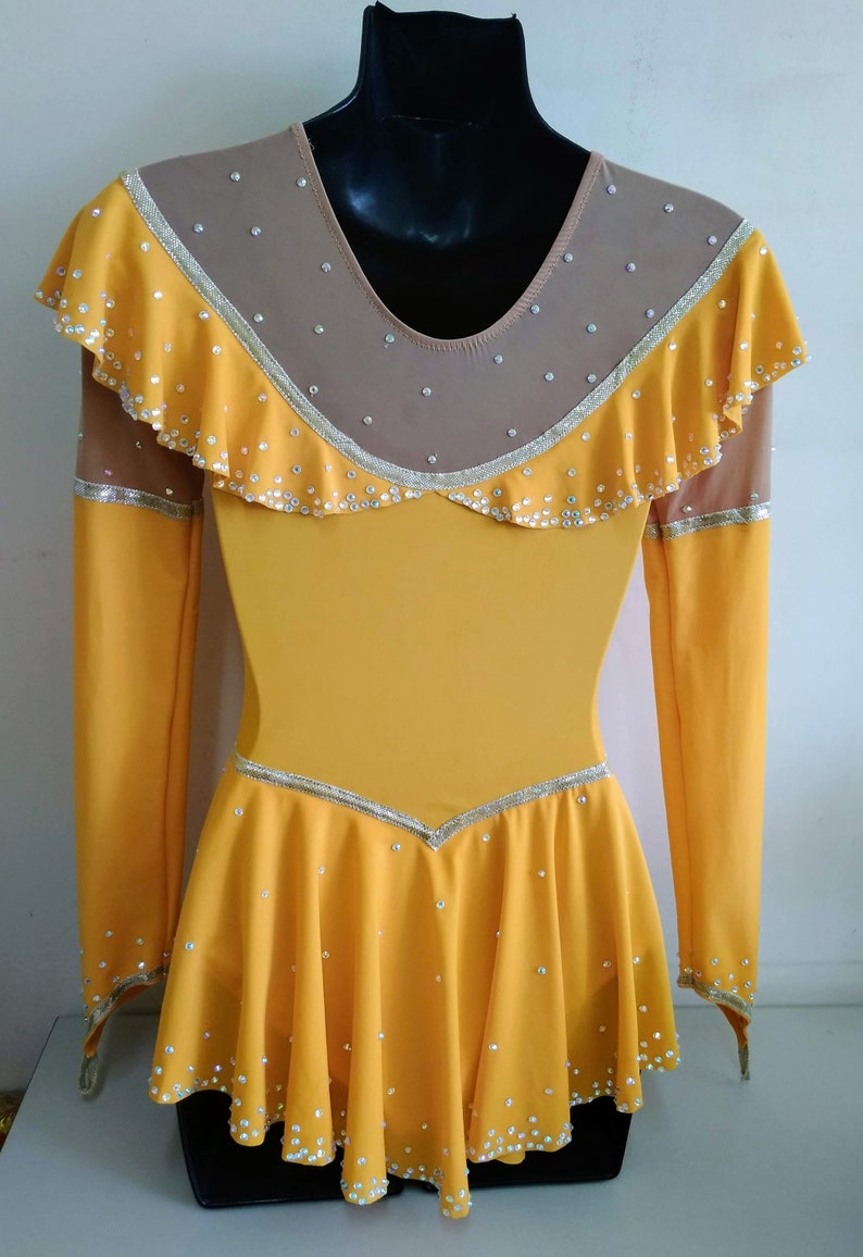 K&ouml;nnte beinhalten: Ein gelbes Eiskunstlaufkleid mit langen &Auml;rmeln und einem R&uuml;schenkragen. Das Kleid ist mit Strasssteinen und einem goldenen Besatz verziert.