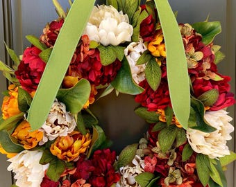 May include: A vibrant floral wreath with a variety of flowers in shades of red, yellow, and cream, accented by green leaves. A wide, light green ribbon is tied at the top, creating a decorative accent. This wreath is perfect for adding a touch of color to any door.