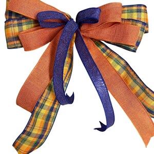 May include: A decorative bow featuring a combination of orange, blue, and plaid ribbons. The bow has a central knot with blue embossed ribbon and long orange and plaid streamers. The plaid ribbon incorporates yellow, orange, and blue.