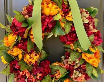 May include: A vibrant floral wreath featuring a mix of red and yellow flowers with green leaves. A light green ribbon is tied at the top for hanging. The wreath is a decorative item, perfect for adding a touch of color to any space.