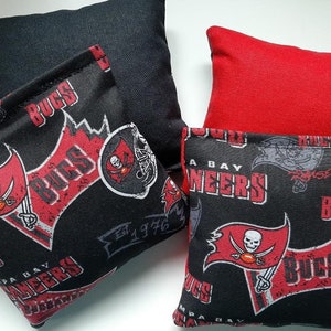 May include: Four bean bags with a Tampa Bay Buccaneers theme. Two bean bags are black with red and white Buccaneers logos and the words "A Bay Buccaneers" and "Est 1976". One bean bag is red and the other is black with the words "Raise the Red" and "Buccaneers".