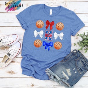 Op de afbeelding: Een blauw T-shirt met een witte letter K, rode, witte en blauwe strikken en basketbals. De tekst "RCJH" staat op het T-shirt.