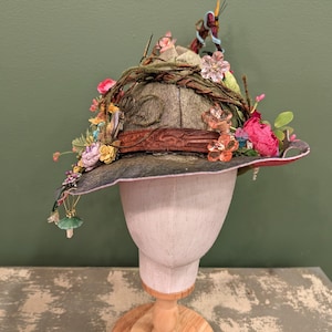 May include: A grey felt hat adorned with a brown leather band, intertwined brown vines, and an assortment of colorful artificial flowers, including pink, yellow, and purple blossoms. Small charms, including a green mushroom, hang from the brim.