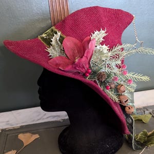 May include: A burgundy witch hat with a wide brim, decorated with a large pink flower, holly, pine needles, berries, and small bells. A silver star hangs from the tip of the hat.