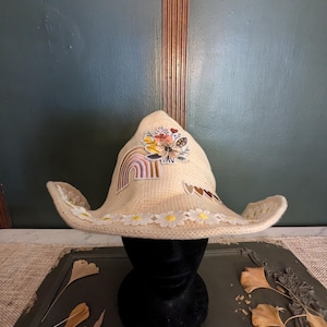 Puede incluir: Un sombrero de vaquero beige con ala ancha, decorado con parches florales y arcoíris. El ala está adornada con encaje blanco y aplicaciones de margaritas. El sombrero se exhibe sobre una cabeza de maniquí negra.