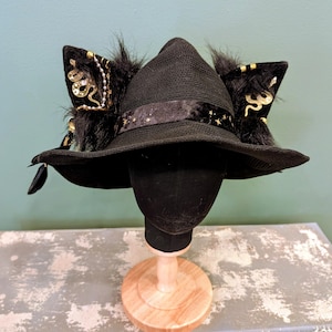 May include: A black wide-brimmed hat featuring two large, upright cat-like ears made of black faux fur. The hat is adorned with a dark ribbon featuring gold star patterns and gold-colored snake embellishments on the ears with small beads.