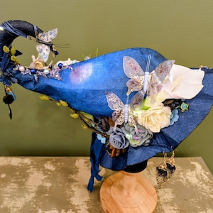 May include: A deep blue pointed hat featuring a curved tip, adorned with translucent butterfly ornaments, white and grey fabric roses, and small green sprigs. The hat includes dangling charms, blue ribbon accents, and delicate beadwork along the crown.
