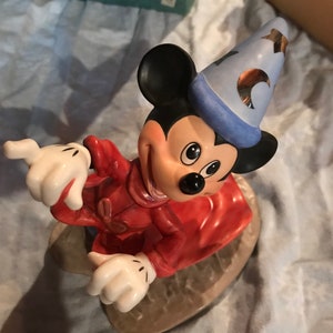 Może przedstawiać: Ceramiczna figurka Myszki Miki przebranej za czarodzieja. Ma na sobie czerwoną szatę, niebieski spiczasty kapelusz z złotymi gwiazdami i białe rękawiczki. Uśmiecha się i ma przebiegły wyraz twarzy.