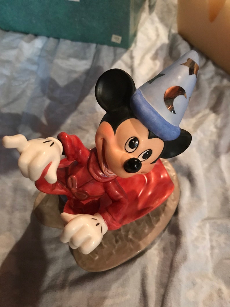 Może przedstawiać: Ceramiczna figurka Myszki Miki przebranej za czarodzieja. Ma na sobie czerwoną szatę, niebieski spiczasty kapelusz z złotymi gwiazdami i białe rękawiczki. Uśmiecha się i ma przebiegły wyraz twarzy.