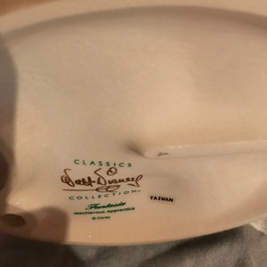 Może przedstawiać: Biała porcelanowa podstawa figurki z tekstem "Classics Walt Disney Collection" i "Fantasia Mischievous Apprentice" w zielonym kolorze. Podstawa zawiera r&oacute;wnież tekst "Taiwan" i symbol praw autorskich Disneya.