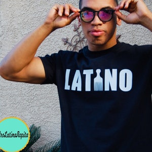 Puede incluir: Una camiseta negra con la palabra "LATINO" impresa en letras blancas. La "I" de "LATINO" est&aacute; reemplazada por una silueta blanca de un peine afro.
