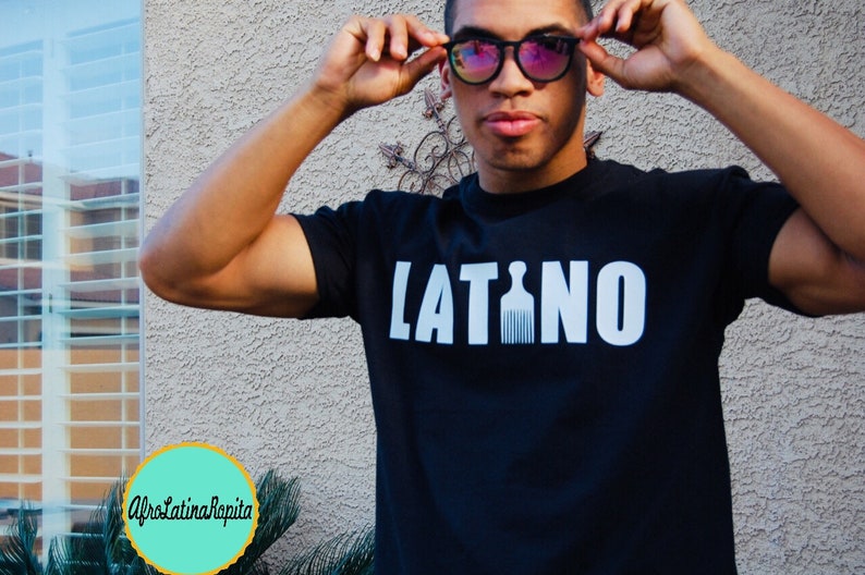 Puede incluir: Una camiseta negra con la palabra "LATINO" impresa en letras blancas. La "I" de "LATINO" est&aacute; reemplazada por una silueta blanca de un peine afro.