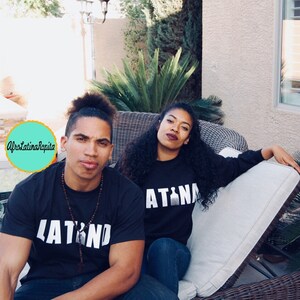 Puede incluir: Una pareja vistiendo camisetas negras a juego con texto blanco. La camiseta del hombre dice "LATINO" y la camiseta de la mujer dice "LATINA". Est&aacute;n sentados en un sof&aacute; de mimbre al aire libre.