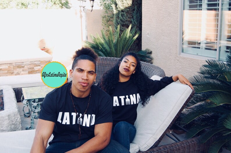 Puede incluir: Una pareja vistiendo camisetas negras a juego con texto blanco. La camiseta del hombre dice "LATINO" y la camiseta de la mujer dice "LATINA". Est&aacute;n sentados en un sof&aacute; de mimbre al aire libre.