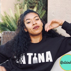 Puede incluir: Una mujer con el pelo largo y negro rizado lleva una sudadera negra con la palabra "LATINA" impresa en letras blancas con un gr&aacute;fico de peine. La sudadera tambi&eacute;n tiene un peque&ntilde;o c&iacute;rculo turquesa con el texto "AfroLatinaRapita" impreso en blanco.