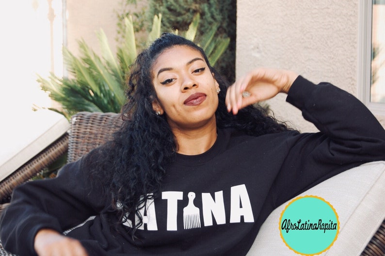 Puede incluir: Una mujer con el pelo largo y negro rizado lleva una sudadera negra con la palabra "LATINA" impresa en letras blancas con un gr&aacute;fico de peine. La sudadera tambi&eacute;n tiene un peque&ntilde;o c&iacute;rculo turquesa con el texto "AfroLatinaRapita" impreso en blanco.
