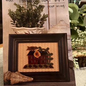 May include: A framed punchneedle embroidery pattern titled "Spring Welcome" by designer Shawn Williams. The pattern features a small house with a sunflower on the roof and a green vine. The pattern is 5 inches by 3 1/2 inches.
