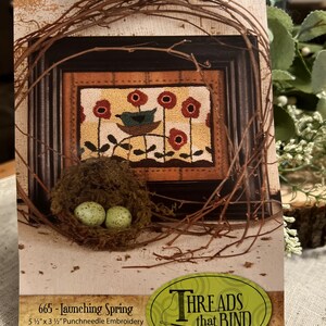 May include: A framed punchneedle embroidery piece titled "Launching Spring" featuring a bird in a nest with red flowers. The embroidery is surrounded by a twig wreath and a nest with two speckled eggs. The dimensions are 5 1/2" x 3 1/2".