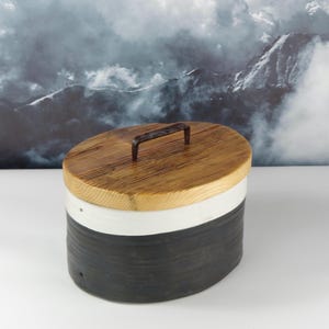 Könnte beinhalten: Ein ovaler Keramikbehälter mit Holzdeckel und Metallgriff. Der Behälter hat einen schwarzen Sockel, einen weißen Mittelteil und einen Deckel aus Naturholz. Der Hintergrund zeigt eine Berglandschaft. Ideal für die Aufbewahrung.