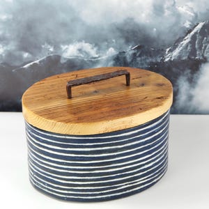 Könnte beinhalten: Ovaler Keramikbehälter mit Holzdeckel und Metallgriff. Der Behälter ist marineblau mit weißen horizontalen Streifen. Der Deckel ist aus hellbraunem Holz. Der Griff ist aus dunkelbraunem Metall.