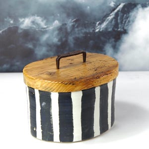 Könnte beinhalten: Ovale Holzbox mit einem gestreiften schwarz-weißen Körper und einem Holzdeckel. Der Deckel hat einen kleinen Metallgriff. Die Box steht vor einer Bergkulisse.