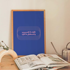 Pu&ograve; includere: Una stampa incorniciata con sfondo blu e testo rosa che recita "نلوم ما نتعب ونحسب ما نكره". La stampa &egrave; appoggiata su un tavolo di legno con un libro e un annaffiatoio.