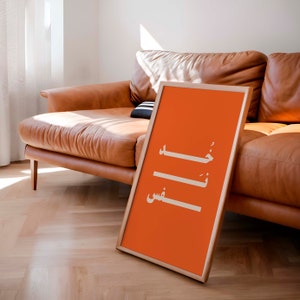 Puede incluir: Un p&oacute;ster naranja enmarcado con caligraf&iacute;a &aacute;rabe blanca que dice "خَلَصْتُ" y "عِشْ" en un sof&aacute; de cuero marr&oacute;n claro.