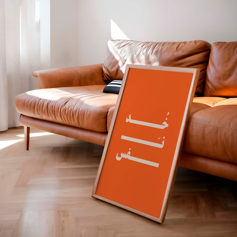 Puede incluir: Un p&oacute;ster naranja enmarcado con caligraf&iacute;a &aacute;rabe blanca que dice "خَلَصْتُ" y "عِشْ" en un sof&aacute; de cuero marr&oacute;n claro.