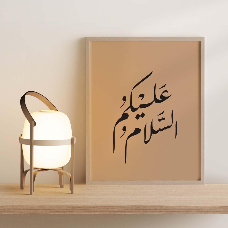 May include: A framed print with a beige background and black Arabic calligraphy that reads "Allah is with you". The print is sitting on a wooden shelf next to a white lamp with a wooden base.