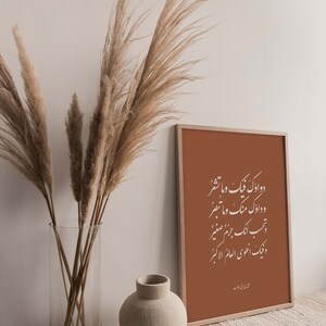 May include: A framed print with a brown background and white Arabic calligraphy. The print is hanging on a wall with a vase of dried pampas grass in front of it. The vase is clear glass and the pampas grass is a light brown color. There is a small, round, beige ceramic vase on the table in front of the print.