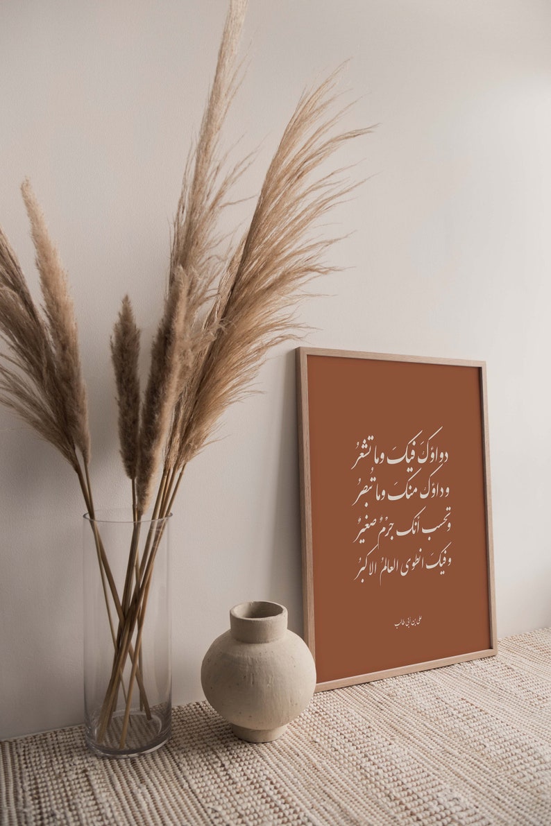 May include: A framed print with a brown background and white Arabic calligraphy. The print is hanging on a wall with a vase of dried pampas grass in front of it. The vase is clear glass and the pampas grass is a light brown color. There is a small, round, beige ceramic vase on the table in front of the print.