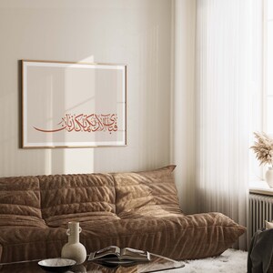 May include: A brown couch with a framed print of Arabic calligraphy in a living room with a white rug and a window with white curtains.