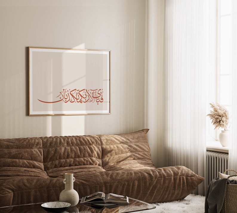May include: A brown couch with a framed print of Arabic calligraphy in a living room with a white rug and a window with white curtains.