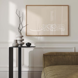 May include: A framed print with a beige background and white Arabic calligraphy that reads "قُلْ هُوَ اللَّهُ أَحَدٌ" (Say, He is Allah, the One). The print is hanging on a white wall above a black side table with a vase and other decorative objects.
