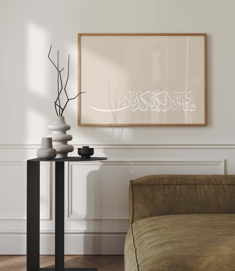 May include: A framed print with a beige background and white Arabic calligraphy that reads "قُلْ هُوَ اللَّهُ أَحَدٌ" (Say, He is Allah, the One). The print is hanging on a white wall above a black side table with a vase and other decorative objects.