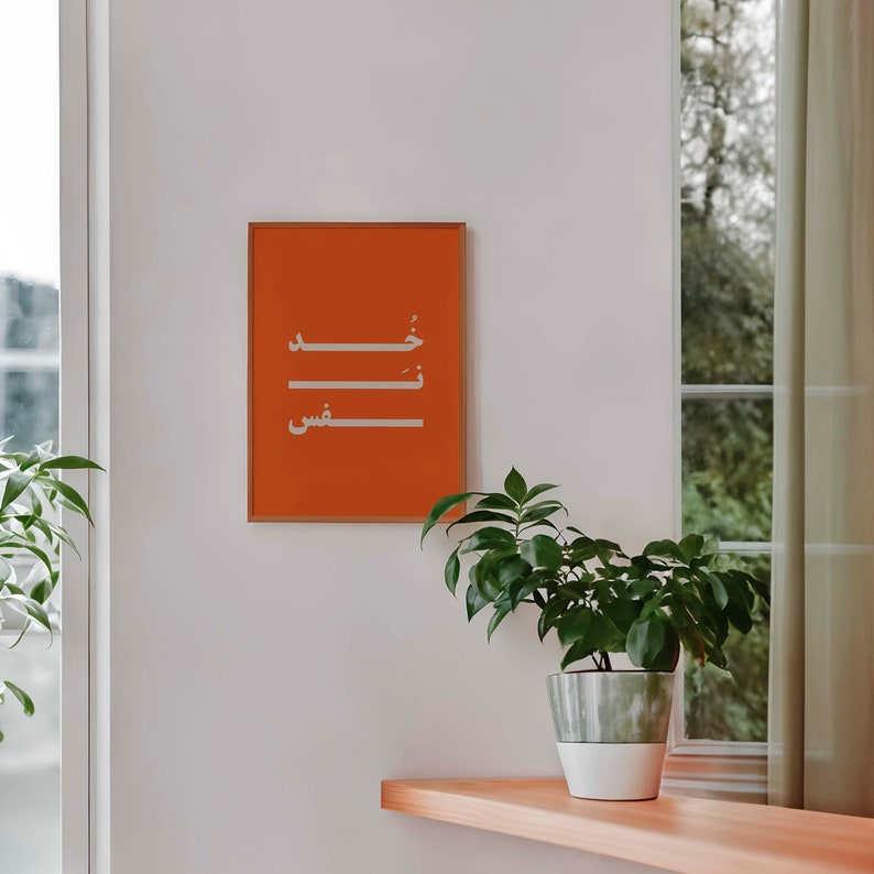 Puede incluir: Una impresi&oacute;n enmarcada con fondo naranja y caligraf&iacute;a &aacute;rabe blanca. La impresi&oacute;n est&aacute; colgada en una pared blanca. Una planta en maceta se encuentra en un estante de madera debajo de la impresi&oacute;n.