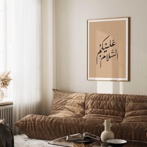 May include: A beige framed print with black Arabic calligraphy on a light brown background. The text reads "عليك السلام" which translates to "Peace be upon you". The print is hanging on a wall above a brown couch.