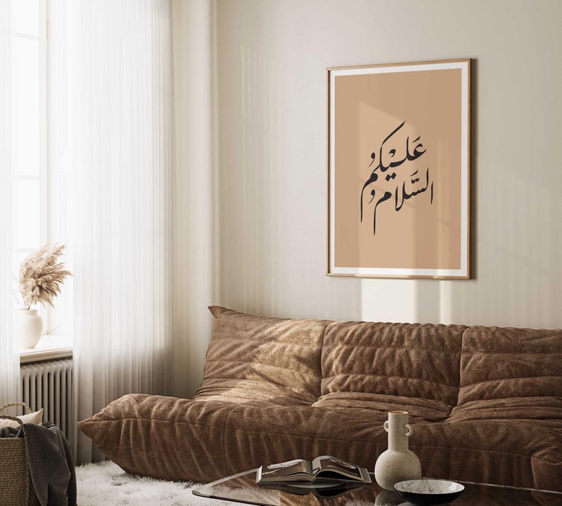 May include: A beige framed print with black Arabic calligraphy on a light brown background. The text reads "عليك السلام" which translates to "Peace be upon you". The print is hanging on a wall above a brown couch.