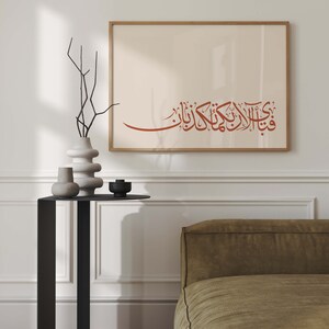 May include: A framed print with a beige background and a red Arabic calligraphy inscription. The print is hanging on a white wall above a black side table with two white vases and a black candle holder. A brown couch is in the background.