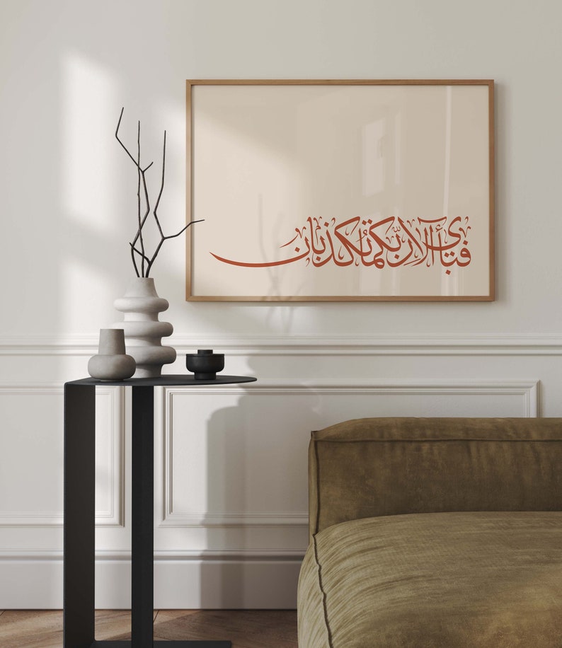 May include: A framed print with a beige background and a red Arabic calligraphy inscription. The print is hanging on a white wall above a black side table with two white vases and a black candle holder. A brown couch is in the background.