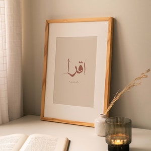 Puede incluir: Una impresi&oacute;n enmarcada con la palabra &aacute;rabe para "leer" en caligraf&iacute;a marr&oacute;n sobre un fondo beige. La palabra est&aacute; rodeada por una l&iacute;nea curva fina. El marco es de madera clara. Un libro, un jarr&oacute;n peque&ntilde;o y un portavelas est&aacute;n sobre una mesa blanca.