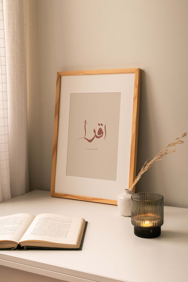 Puede incluir: Una impresi&oacute;n enmarcada con la palabra &aacute;rabe para "leer" en caligraf&iacute;a marr&oacute;n sobre un fondo beige. La palabra est&aacute; rodeada por una l&iacute;nea curva fina. El marco es de madera clara. Un libro, un jarr&oacute;n peque&ntilde;o y un portavelas est&aacute;n sobre una mesa blanca.