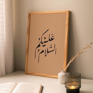 May include: A framed print with a brown background and black Arabic calligraphy that reads "Allah is with you". The print is on a white table with a book, a small white vase, and a candle holder.