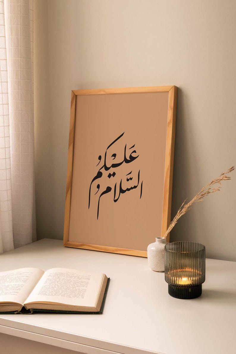 May include: A framed print with a brown background and black Arabic calligraphy that reads "Allah is with you". The print is on a white table with a book, a small white vase, and a candle holder.