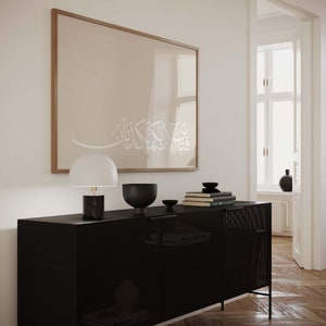May include: A black metal cabinet with a grid pattern on the doors. The cabinet has a white lamp with a black base and a black bowl on top. There is a framed print with Arabic calligraphy on the wall behind the cabinet.