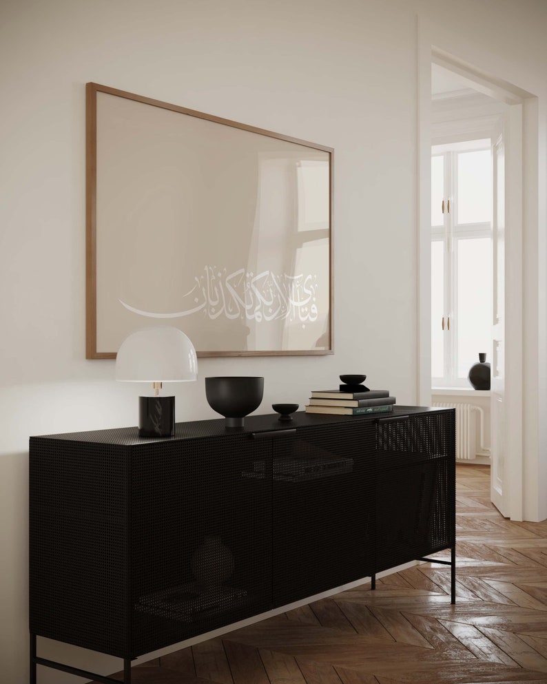 May include: A black metal cabinet with a grid pattern on the doors. The cabinet has a white lamp with a black base and a black bowl on top. There is a framed print with Arabic calligraphy on the wall behind the cabinet.